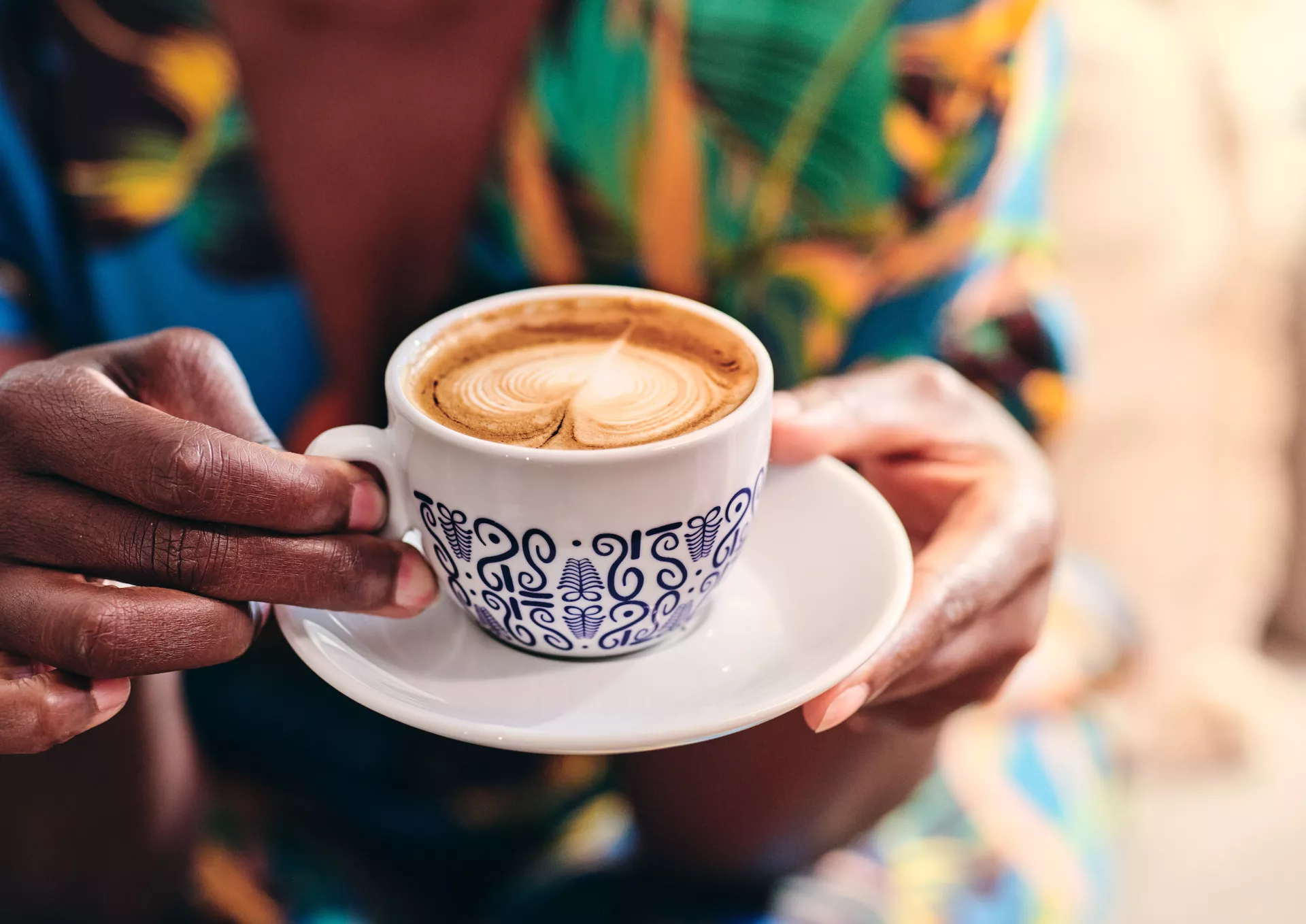 Eine Person hält eine Tasse Cappuccino mit Milchschaum auf einer Untertasse in den Händen.